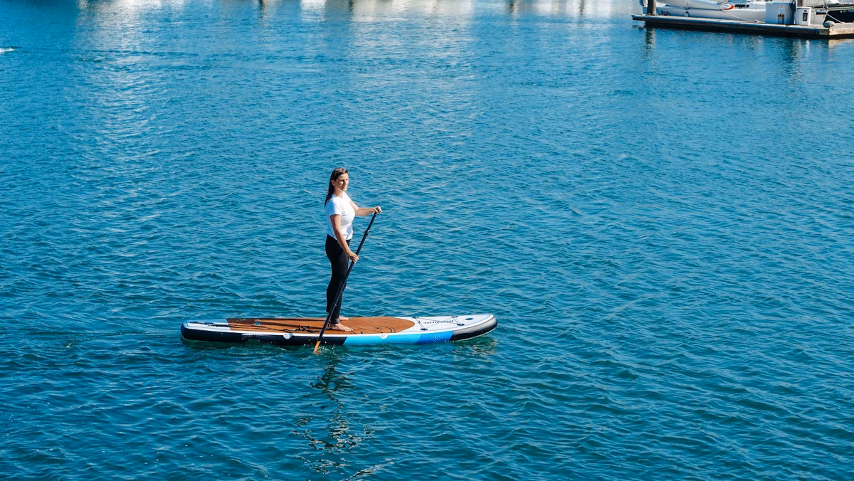 Home 1 A woman standing and paddling on a wood-deck all-around inflatable stand-up paddle board on calm blue water