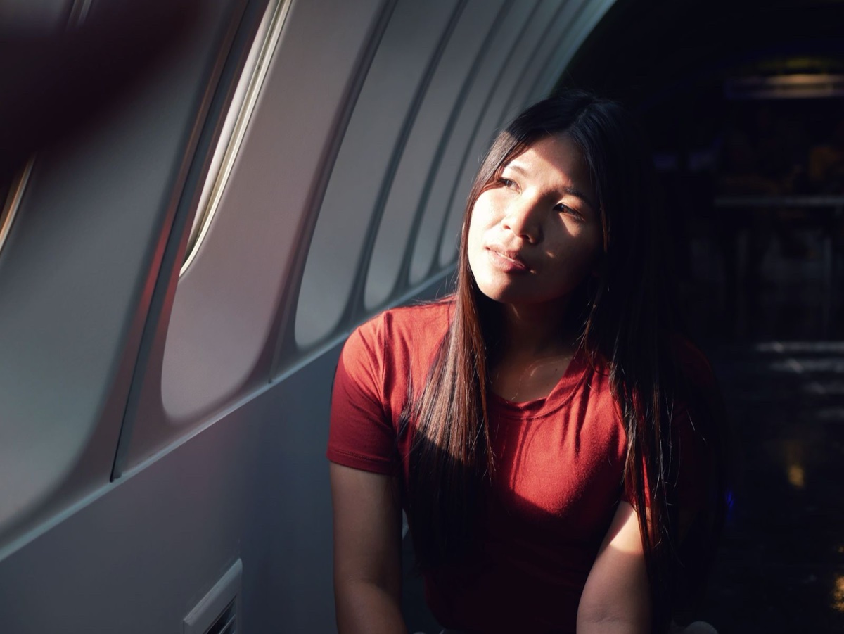 A woman in a red shirt sits in an airplane seat with afternoon sun falling across her face through the cabin window
