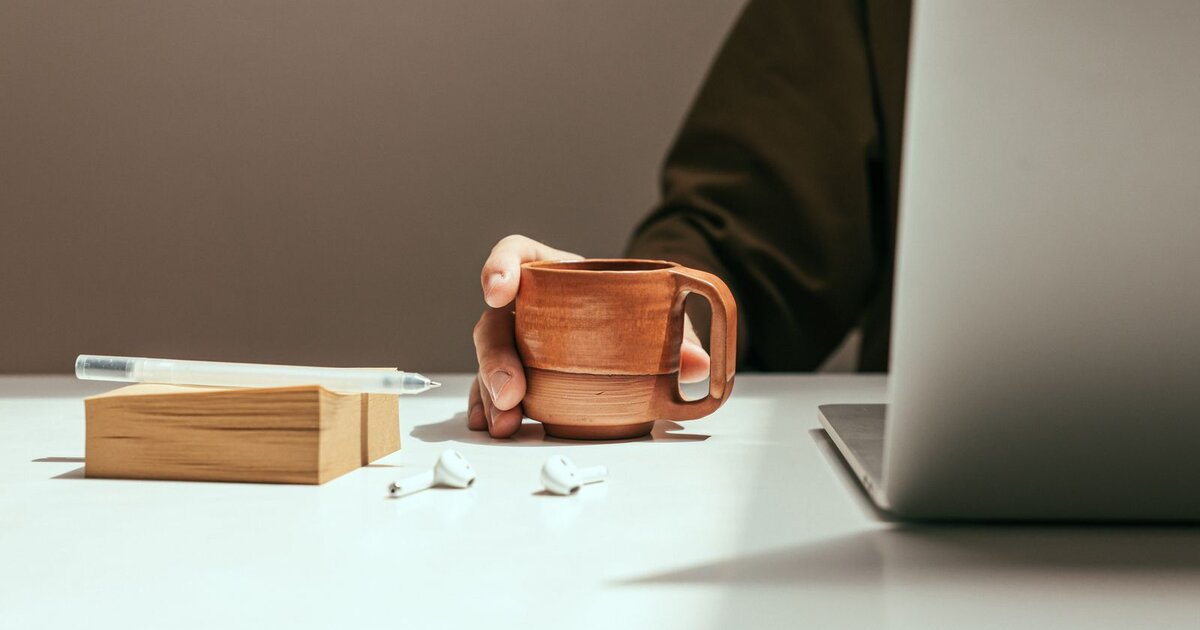 A hand resting on a terracotta coffee mug on a white desk beside a laptop, sticky notes, and wireless earbuds